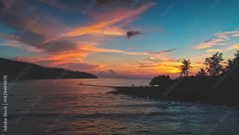 Orange, pink and blue sunset sky over Koh Ma Beach in Koh Phangan, Thailand, with tourists walking along the sandbar at low tide, connecting Koh Ma to the main island