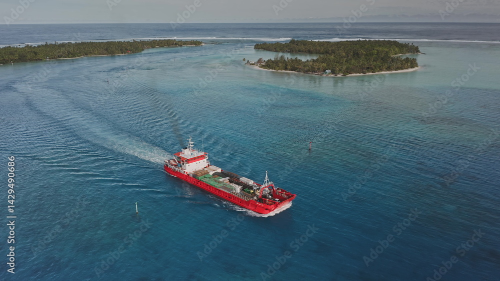 Red cargo ship sailing through turquoise waters near a tropical island, leaving a white trail against a backdrop of sunny skies dotted with a few clouds