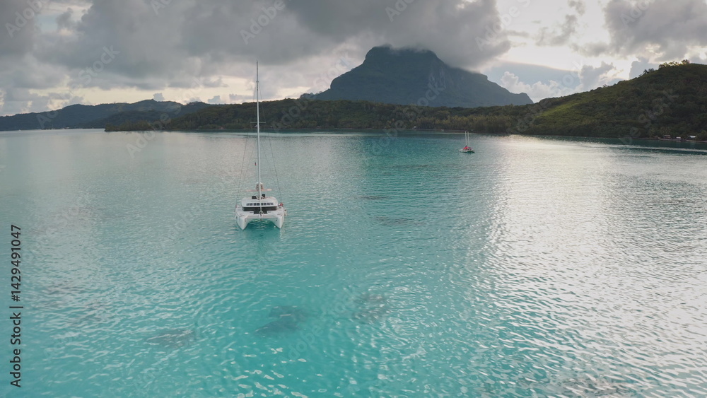 Bora Bora discovery and travel: white catamaran sailing in tranquil turquoise water with mount Otemanu in background under cloudy sky, creating serene tropical paradise view. Remote wild nature scene