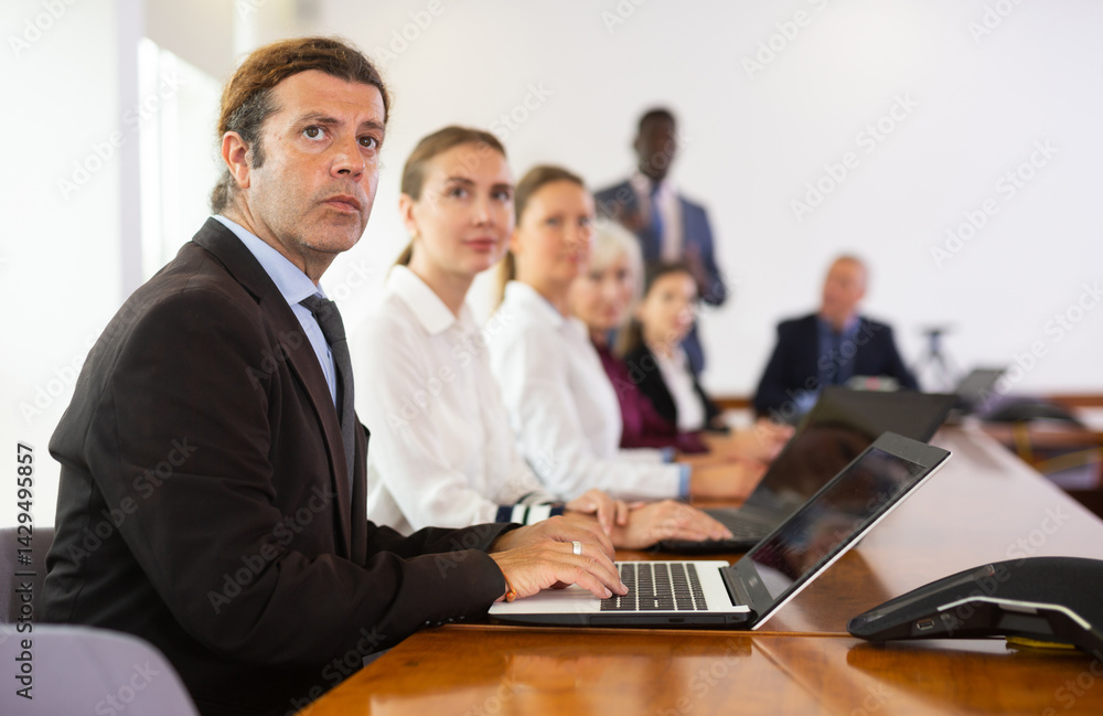 Serious middle-aged white brunette businessman sitting among colleagues at conference table in meeting room, intently listening to presentation and taking some notes on laptop during corporate