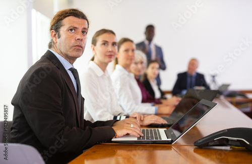 Obraz na płótnie Serious middle-aged white brunette businessman sitting among colleagues at conference table in meeting room, intently listening to presentation and taking some notes on laptop during corporate w salonie