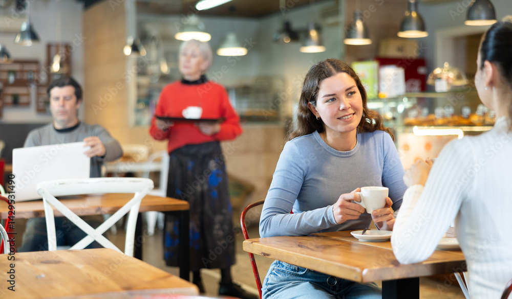 Girl in cafe sits opposite female companion, has nice conversation and discusses news of day. Cozy atmosphere of cafe, meeting place for friends, business partners, colleagues. Girl in cafe sits opposite female companion, has nice conversation and discusses news of day. Cozy atmosphere of cafe, meeting place for friends, business partners, colleagues.