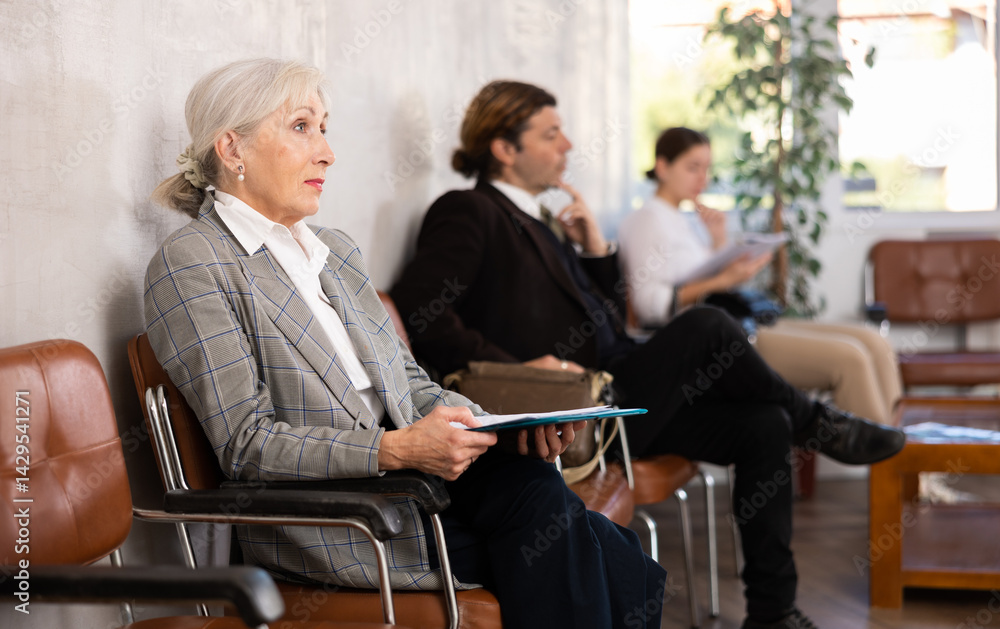 Elderly woman in business suit holds folder with documents sitting on chair in office
