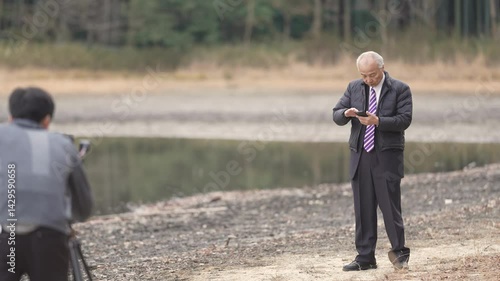 There is a lot of greenery in the bamboo forest pond at noon in spring in Japan. Japanese men in the late 70s stood in suits and down jackets. His son is taking a commemorative photo.