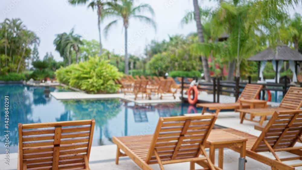 Blurred view of luxurious tropical resort with wooden lounge chairs by a serene pool surrounded by lush greenery and palm trees under a soft sky. Blurred view of luxurious tropical resort with wooden lounge chairs by a serene pool surrounded by lush greenery and palm trees under a soft sky.