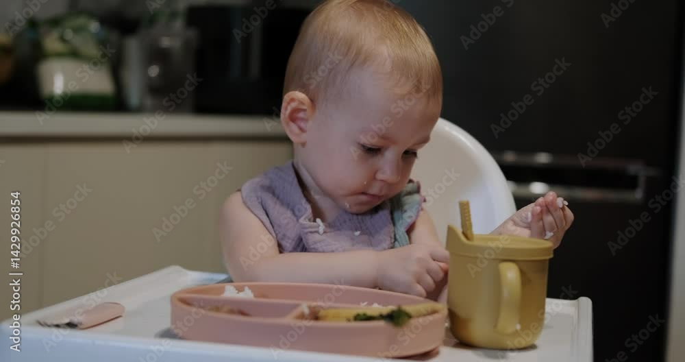 A little toddler girl sits in a high chair and eats her lunch. The baby is learning to eat with a fork, but she picks up rice with her hand and puts it in her mouth. She plays with food and has fun. A little toddler girl sits in a high chair and eats her lunch. The baby is learning to eat with a fork, but she picks up rice with her hand and puts it in her mouth. She plays with food and has fun.