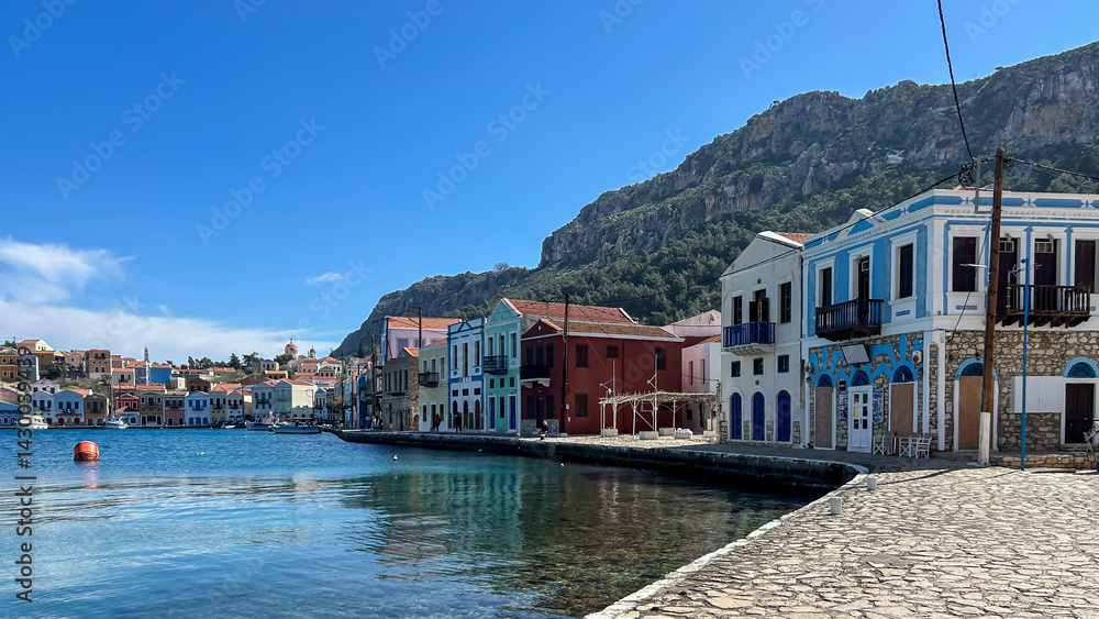 Obraz premium Colorful Waterfront Houses In Kastellorizo, Greece, Lined Along A Calm Harbor With Mountain Backdrop, Bright Sunny Day, Concept Of Mediterranean Travel, Real Estate, Summer Vacation, Tourism Promotion