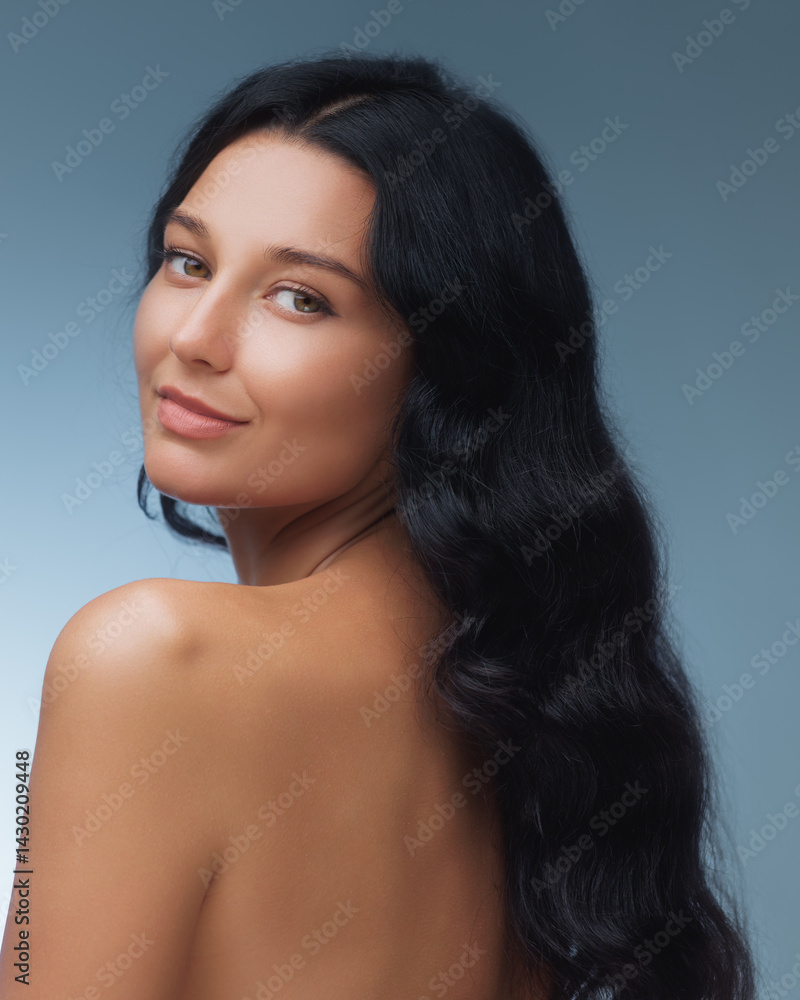A stunning woman with long black hair gazes over her shoulder, highlighting her smooth skin and natural beauty against a soft blue background. Ideal for beauty, skincare, and wellness themes.