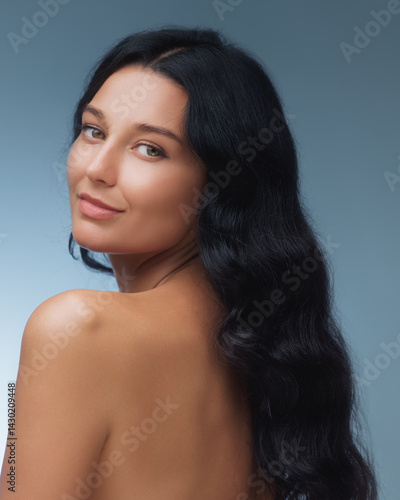 Obraz na płótnie A stunning woman with long black hair gazes over her shoulder, highlighting her smooth skin and natural beauty against a soft blue background. Ideal for beauty, skincare, and wellness themes. w salonie