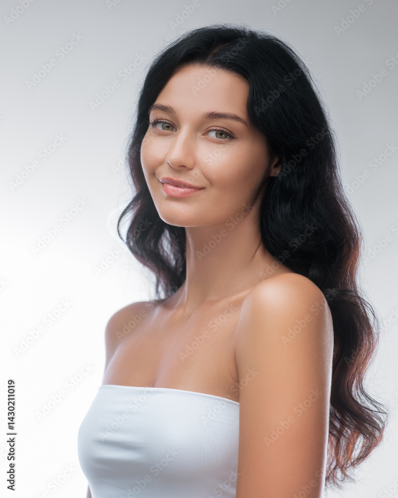 Portrait of a young Caucasian woman with long dark hair and smooth, glowing skin, wearing minimal makeup and a strapless top. Ideal for beauty, skincare, and wellness concepts.