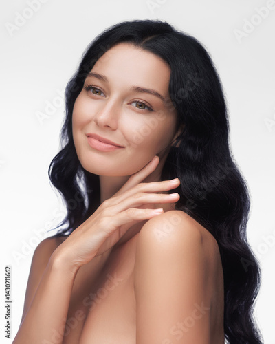 Obraz na płótnie A stunning woman with long black hair and a gentle smile touches her face, showcasing her flawless skin against a white background. Ideal for beauty, skincare, and self-care themes. w salonie