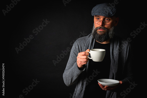 Obraz na płótnie Stylish bearded man in cap and glasses holding coffee cup and saucer on dark background. Thoughtful expression, cozy casual wear, modern vintage atmosphere, morning tea drinking w salonie
