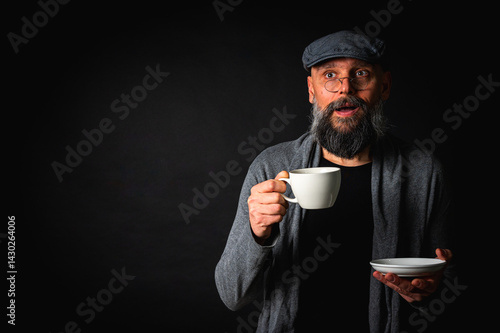 Obraz na płótnie Stylish bearded man in cap and glasses holding coffee cup and saucer on dark background. Thoughtful expression, cozy casual wear, modern vintage atmosphere, morning tea drinking w salonie