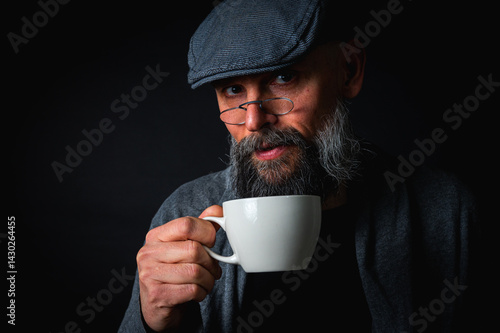 Obraz na płótnie Stylish bearded man in cap and glasses holding coffee cup and saucer on dark background. Thoughtful expression, cozy casual wear, modern vintage atmosphere, morning tea drinking w salonie