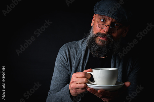 Obraz na płótnie Stylish bearded man in cap and glasses holding coffee cup and saucer on dark background. Thoughtful expression, cozy casual wear, modern vintage atmosphere, morning tea drinking w salonie