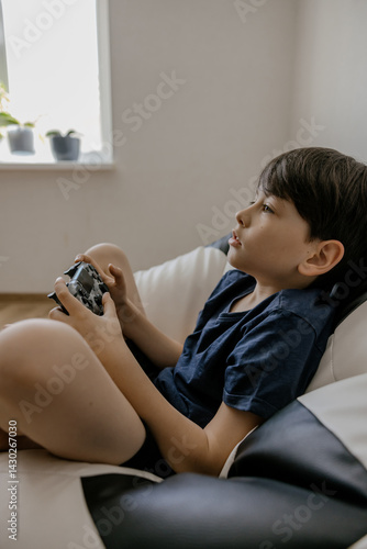 A happy young boy enjoying a video game while sitting in a cozy black and white bean bag chair. He is holding a wireless gaming controller and smiling with excitement. The indoor scene conveys leisure
