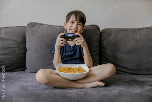 A focused young boy playing video games on a grey couch at home. He is holding a wireless gaming controller and has a bowl of potato chips on his lap. The living room setting includes modern decor 