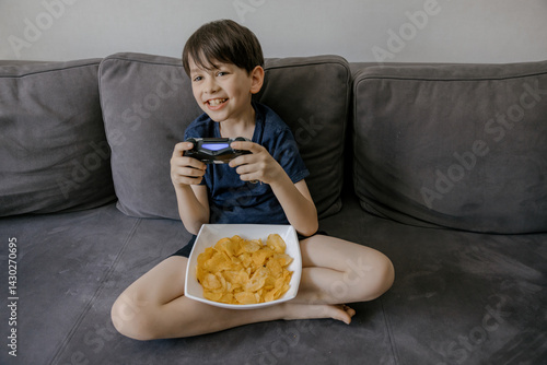 A focused young boy playing video games on a grey couch at home. He is holding a wireless gaming controller and has a bowl of potato chips on his lap. The living room setting includes modern decor 