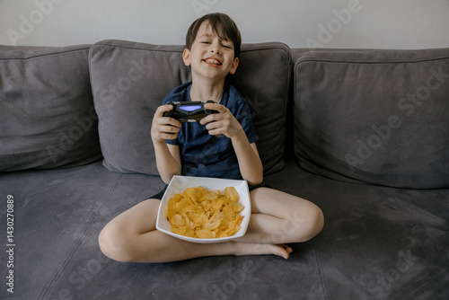 A focused young boy playing video games on a grey couch at home. He is holding a wireless gaming controller and has a bowl of potato chips on his lap. The living room setting includes modern decor 