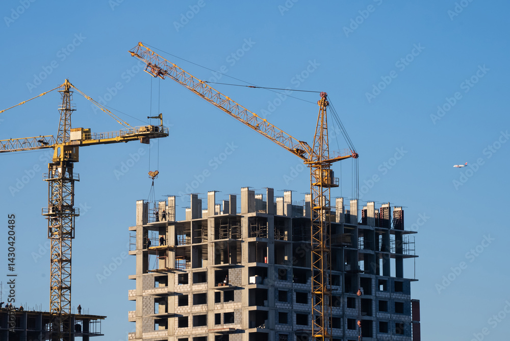 Two construction cranes working on building new modern residential apartment building under clear blue sky, representing urban development and growth Two construction cranes working on building new modern residential apartment building under clear blue sky, representing urban development and growth