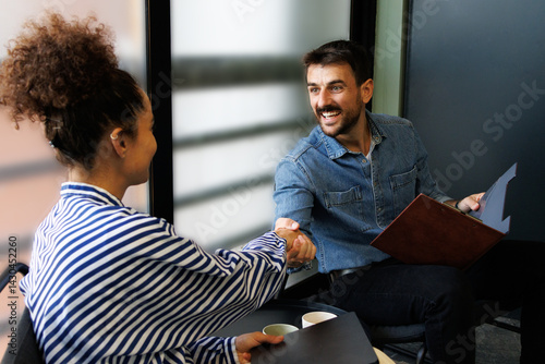 A cheerful business meeting between a man and a woman in a modern office setting. The man is smiling while shaking hands with the woman, who is wearing a striped shirt. 