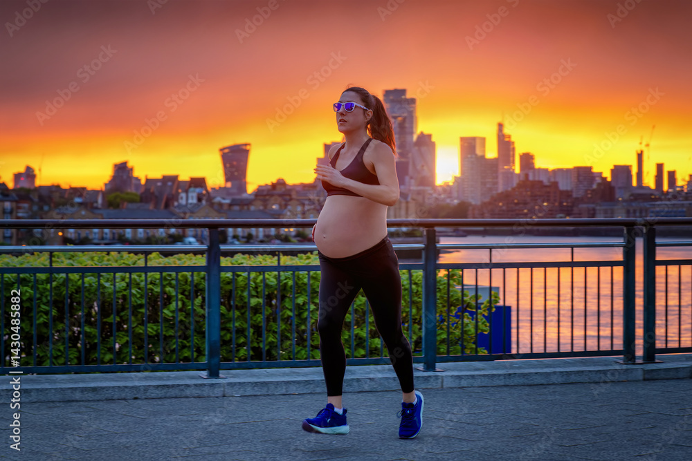 A pregnant woman in sports clothing running for exercise in front of an urban skyline during a fiery sunset