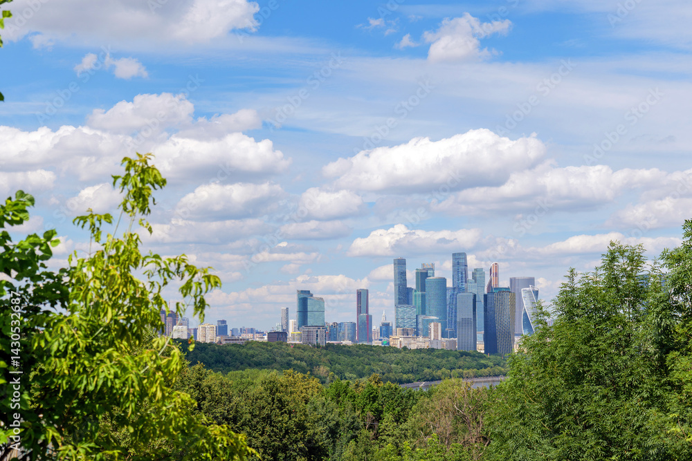 Fototapeta premium Breathtaking view of Moscow skyline from Vorobyovy Gory, with Moscow River, green trees, and clouds in the blue sky. The Moscow skyline dominates the horizon, creating a stunning scene