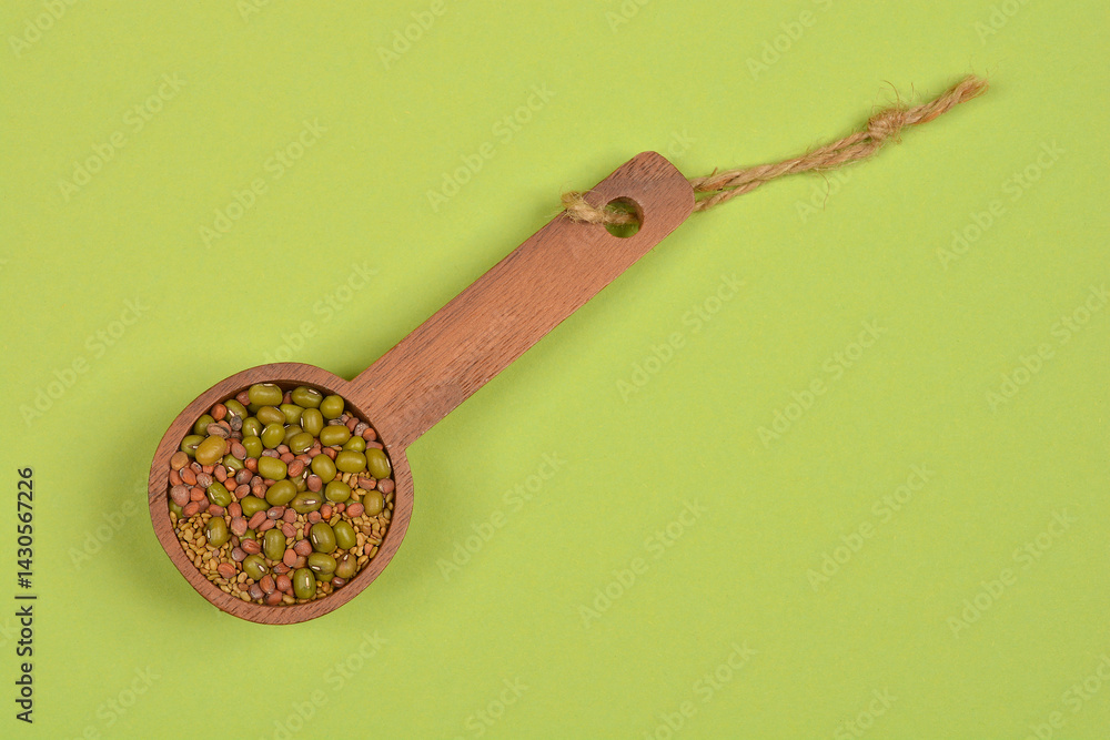 A wooden spoon holds a mix of green mung beans, tan alfalfa seeds, and reddish radish seeds against a solid light green background