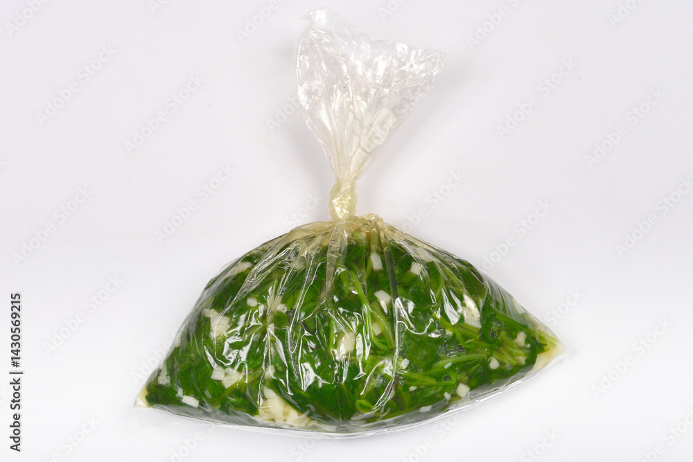 Cooked radish leaves with garlic are sealed in a clear plastic bag on a light gray surface, captured from above