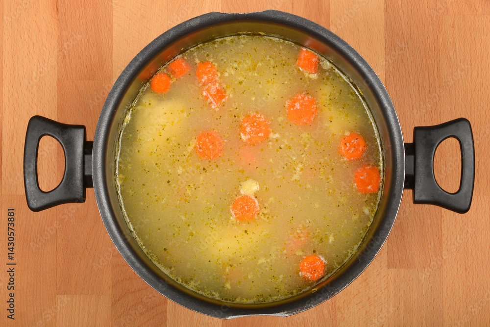 An overhead view shows a pot of clear, light-colored soup with several pale yellow semolina dumplings and bright orange carrot slices, sprinkled with herbs, on a wooden surface