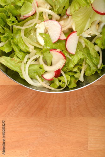 An overhead shot shows a stainless steel bowl filled with a vibrant spring salad of crisp green lettuce, thinly sliced white onions, and red and white radish slices on a wooden surface