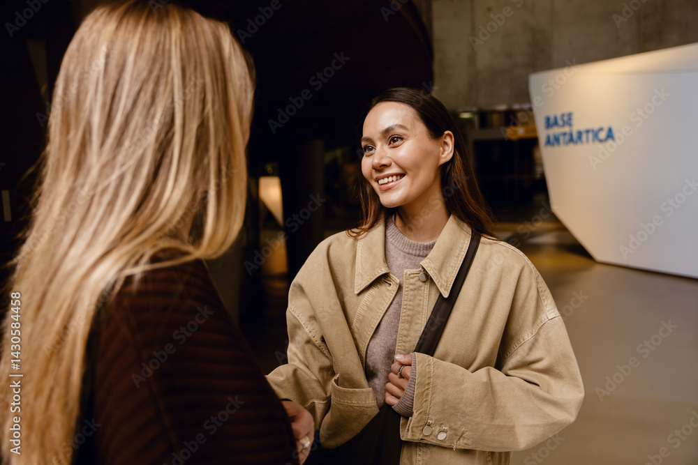 Two young women engage in friendly conversation indoors near a modern exhibit marked