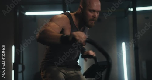 Close up of a male athlete with a shaved head in a tank top doing an intense workout and fast movement on an exercise bike in a modern gym