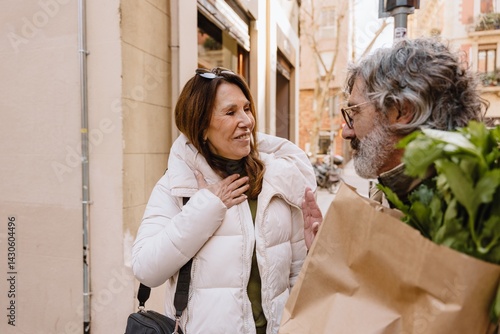 An elderly White couple is talking in the street, gesticulating vigorously. The man holds a shopping bag in his hand and turns to the smiling woman. A concept of routine, bonding, and relationship.