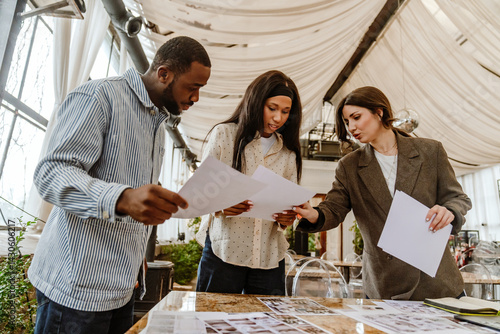A young Black groom and bride are discussing printed materials with a young White female wedding planner in a decorated event venue filled with natural light and draped fabric ceiling.