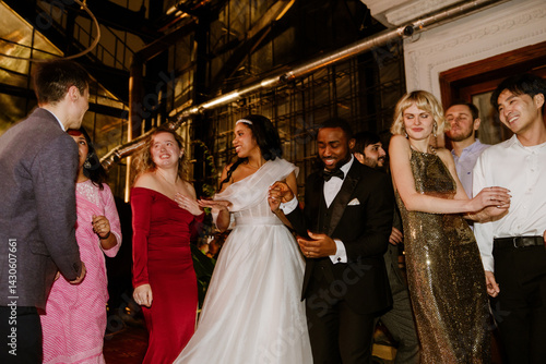Obraz na plátně A multicultural group of adult wedding guests dances indoors with the bride in a white gown and the groom in a black suit during a joyful evening wedding celebration filled with formal outfits