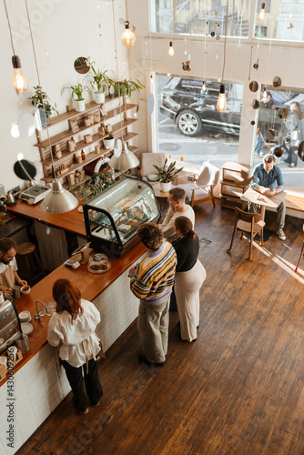 Adults inside a modern cafe with wooden floors and large windows, some standing in line to order from the barista at the counter while one man sits by the window enjoying a drink.