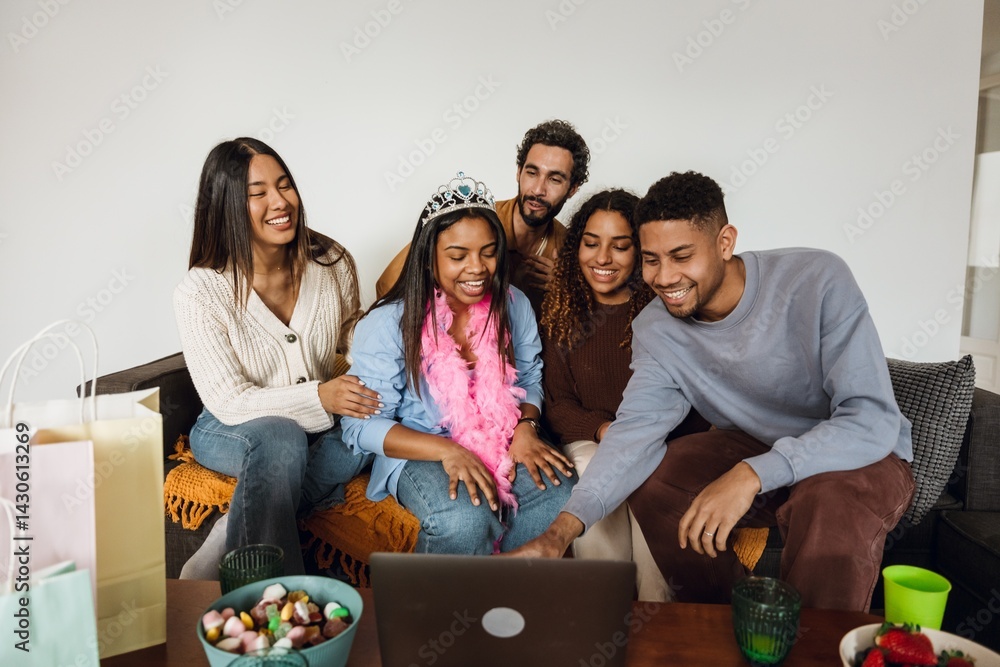 Five multiethnic friends laugh together while looking at a laptop on a sofa. An Indian woman wears a birthday tiara and boa as they enjoy a shared moment watching something funny.