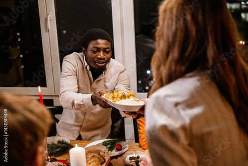 A Black man in his 30s, wearing a white button-up shirt, passing a plate of food to his white-skinned female friend as they're having Christmas dinner with her family, with windows in the background