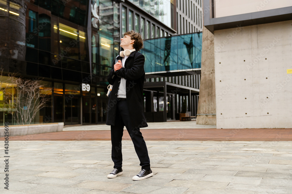 Fototapeta premium In the afternoon in a square with office buildings, a smiling and handsome dark-haired European man in his early 30s in glasses with white headphones in a black coat stands and looks up.