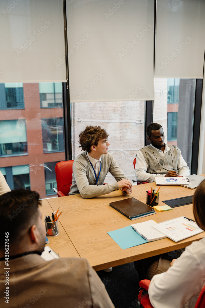 Two young male interns in their late 20s, a White man, and a Black man, sitting among their colleagues at a table in an office meeting room, with windows around them, listening to someone talk