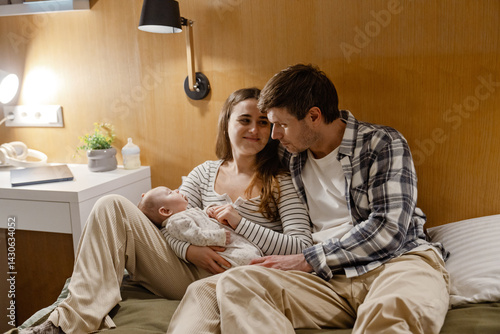 Fotografie A dark-haired European father in his early 30s and a smiling dark-haired adult mother in a striped sweater holding their newborn baby in her arms and enjoying while lying on the bed in the bedroom