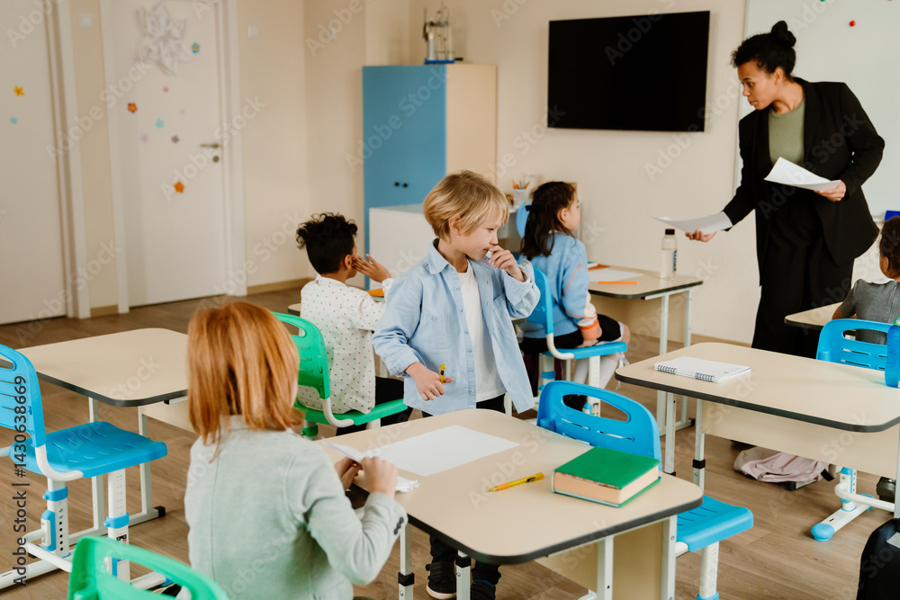 Obraz premium A Black female teacher in her 30s standing in a classroom in front of a multiethnic group of school-aged children sitting at desks during a lesson, a White school-aged boy standing with a pen
