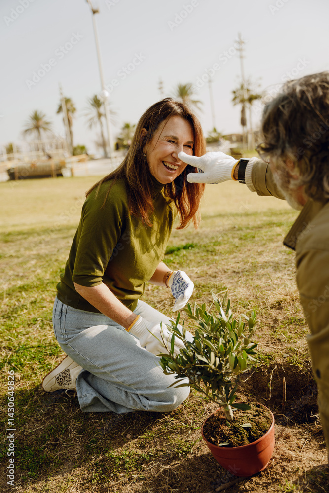 Fototapeta premium An elderly White couple plants outdoors. The smiling woman kneels by a small tree sapling while the man playfully touches her nose. They enjoy a lighthearted moment during their gardening.