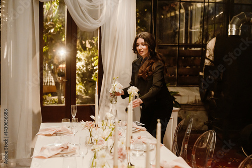 Cuadro en lienzo An adult White woman with long dark hair, dressed in black, carefully places white flowers in small glass vases, arranging centerpieces on a dining table for a wedding in a dimly lit venue