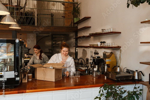 In the afternoon, a happy dark-haired female coffee shop owner in her early 30s in a white shirt is putting glasses out of a box while a red-haired adult friend is cleaning up in the background.