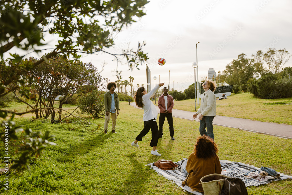 A group of four adult friends aged 25, tossing a volleyball around and having fun as they stand on a grassy field in a park, with their female friend sitting on a blanket beside them and watching
