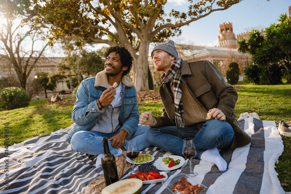 A loving gay couple of a White man and his Black husband, who are in their 30s, sitting on a blanket in the park, having a picnic date and looking off somewhere with a happy smile on their faces