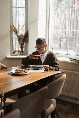 An Asian man in his 70s with dark hair, in glasses, a brown cardigan, and a white shirt, sits at a wooden dining table at home near a window, drinking tea from a cup and flipping through a photo album