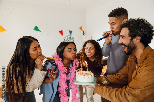 A multiethnic group of friends celebrates a birthday. A young Indian woman wearing a tiara blows out candles on a cake held by a Latino man, surrounded by smiling Asian and Black friends.